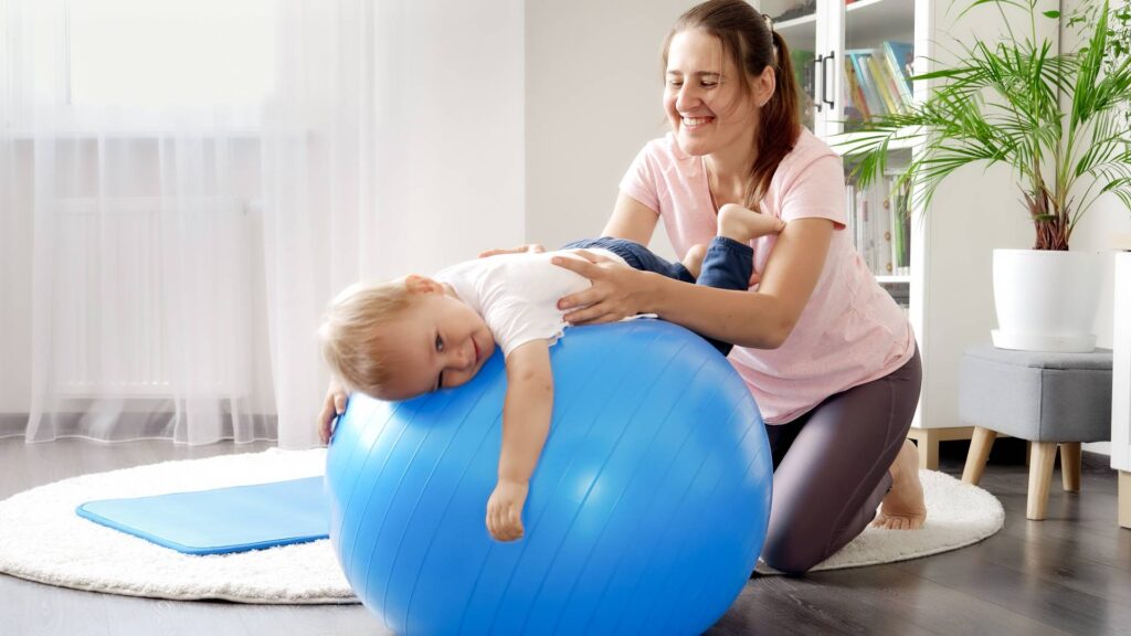 Smiling mother and baby doing fitness exercises on fitball at home. Concept of child healthcare, kids sports and yoga.