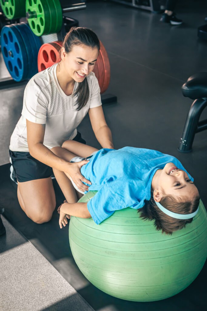 Cute child girl stretching on pilates fitness ball with mom in gym.