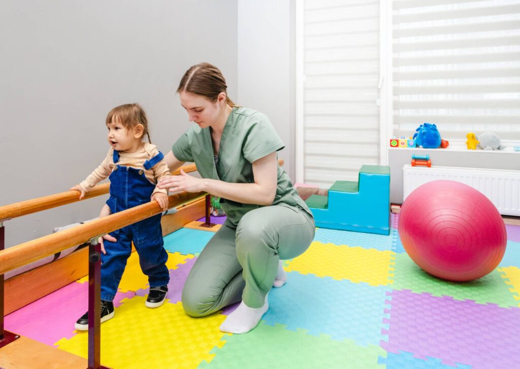 Little girl is assisted by therapist in physical rehabilitation exercises in bright, playful therapy environment. Handrails for restoring walking skills will help in rehabilitation of child